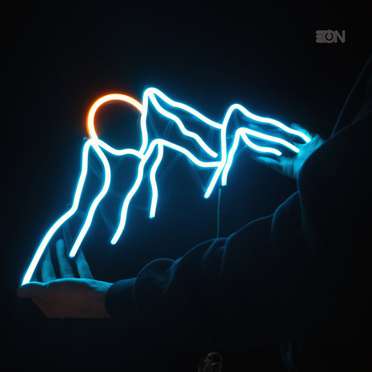 Insegna neon LED tenuta tra le mani. Disegno di una montagna con il sole che tramonta, base 3D nera, luce azzurra e gialla oro.